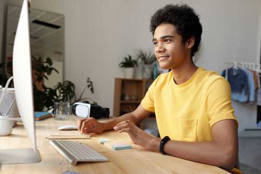 Junger Mann in gelbem T-Shirt sitzt vorm Computer und schaut auf den Bildschirm