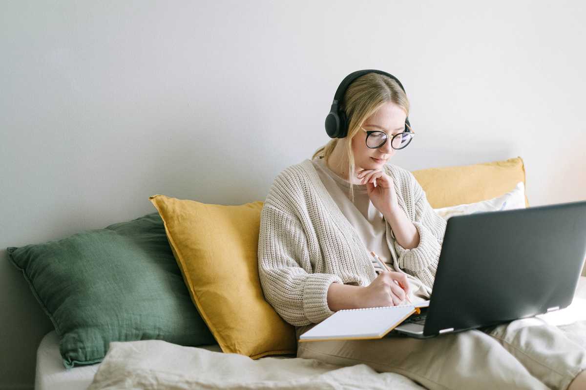 EIne junge Frau mit Kopfhörern sitzt auf einem Bett, hat ihren Laptop auf dem Schoß und schreibt etwas in ein Notizbuch