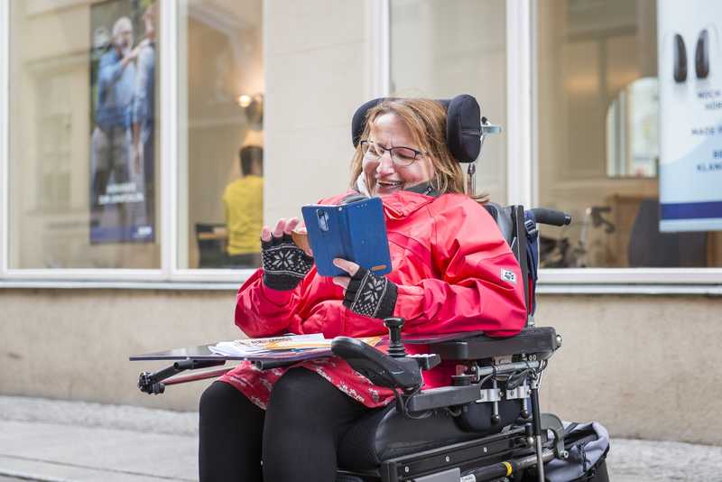 eine Frau mit roter Jacke sitz in einem Rollstuhl und tippt auf dem Handy